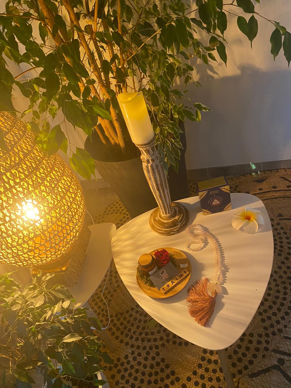 Bougie sur un chandelier en bois cérusé, fleur de frangipanier et mala en quartz rose au pied d'un ficus